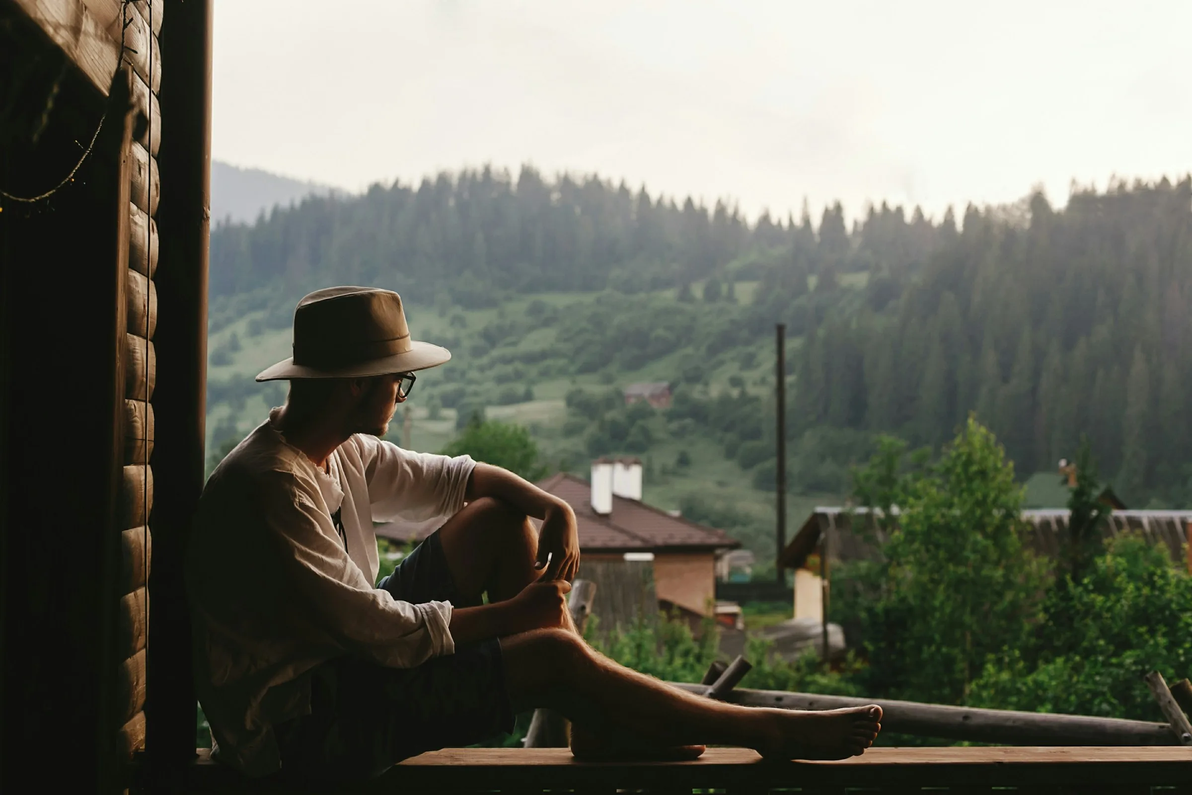 Hombre con sombrero sentado en un balcón de madera contemplando un paisaje de montañas y pinos, representando la introspección y el propósito en las rutas de True Ventour.